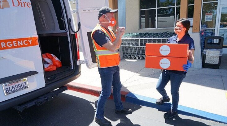 Male worker/driver in a construction vest and mask greeting a female worker, also masked, carrying two boxes to be put in the back of a white van.