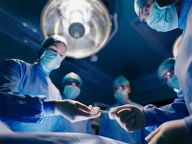 Doctors in scrubs, masks, and eye protection in a surgery room handing one another tools under a bright light