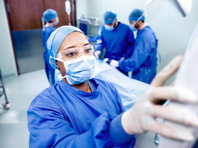 Female doctor in scrubs, mask, eye protection up close typing into a medical device while three other doctors work on a patient in the background that is blurred. 