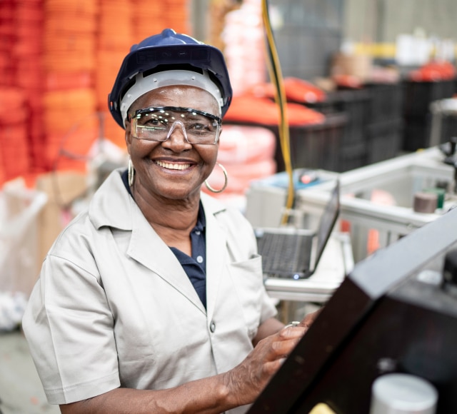 Female factory worker smiling, wearing a hard hat and protective glasses.