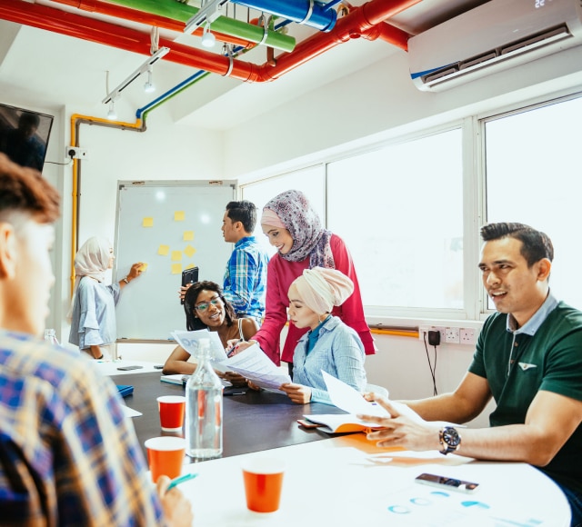 Diverse group of young employees working together in a boardroom setting, sitting at the conference table, writing on the white board and discussing.