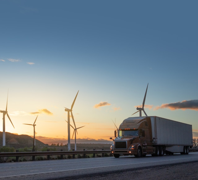 Truck driving across the highway during sunset, with wind turbines in the background with sunshine.